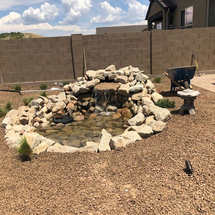 A small rock waterfall and pond in a backyard, surrounded by gravel and a stone wall.