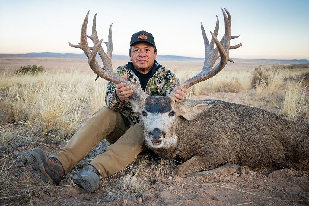 Man in camouflage holds a large buck with massive antlers in a field; desert backdrop.