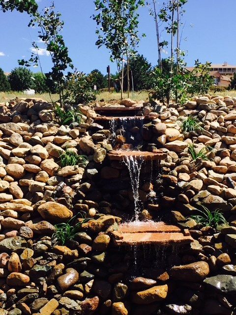 Waterfall cascading over stacked rock ledges, surrounded by rocks and vegetation, under a sunny sky.