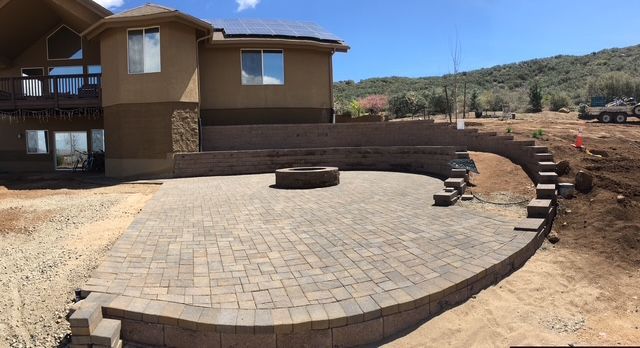 Stone patio with fire pit and retaining walls, next to a two-story house, sunny day.