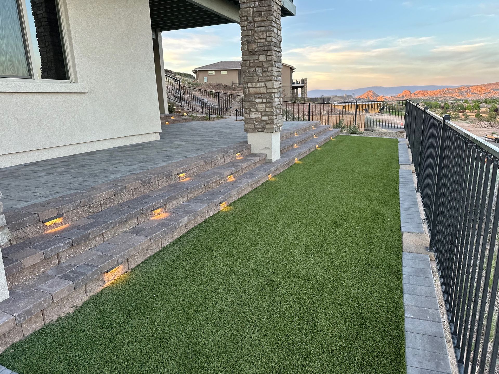 Stone steps with built-in lights leading to a patio with green turf alongside a black fence.