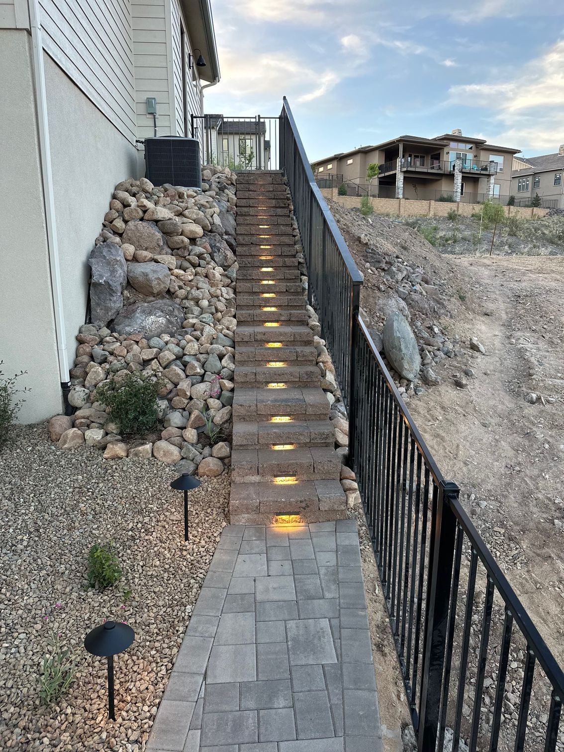 Stone steps with built-in lights ascend alongside a rocky hill and black railing.