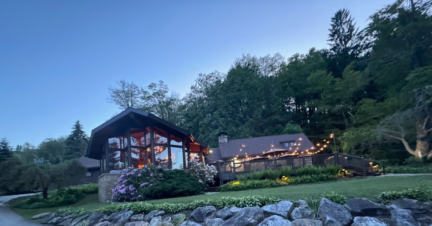 A house at dusk with large windows, string lights, and a stone retaining wall. Set against a treed hill.