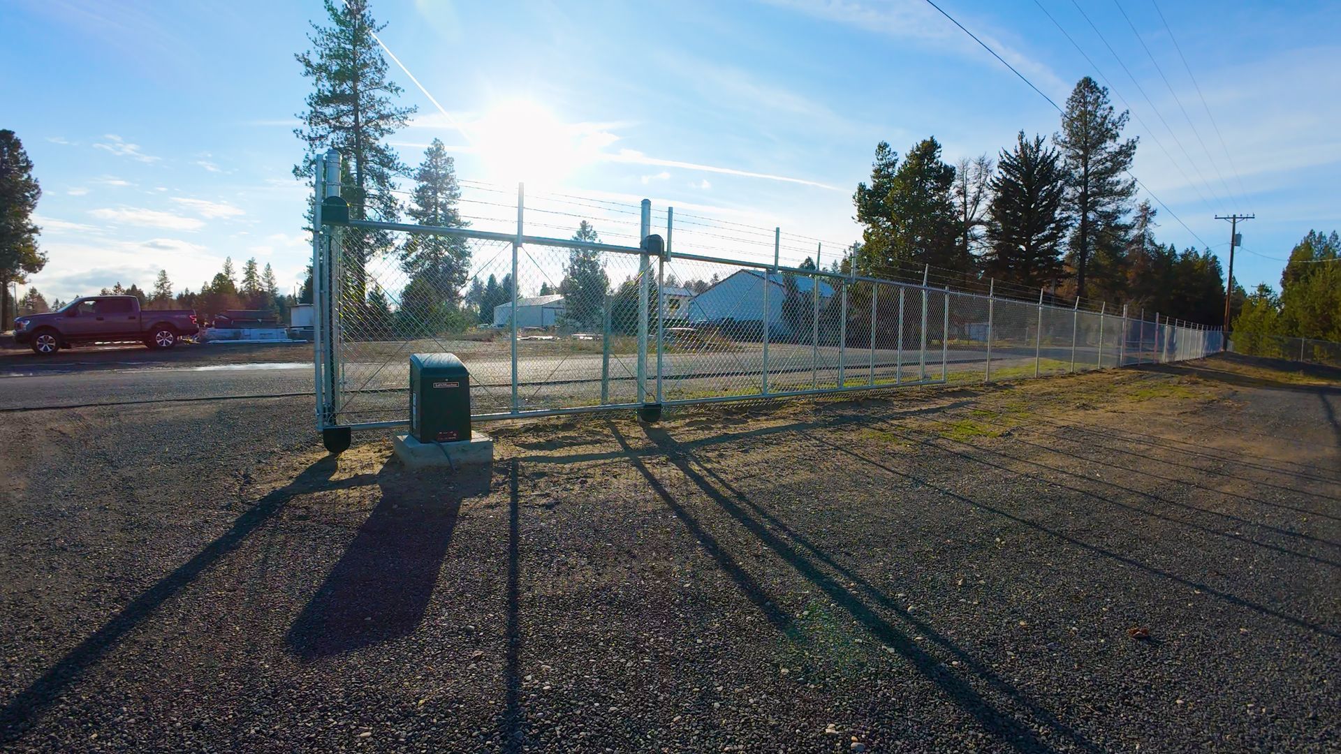 Chain-link fence surrounding a building; a pickup truck and trees are visible in the distance under a bright sky.
