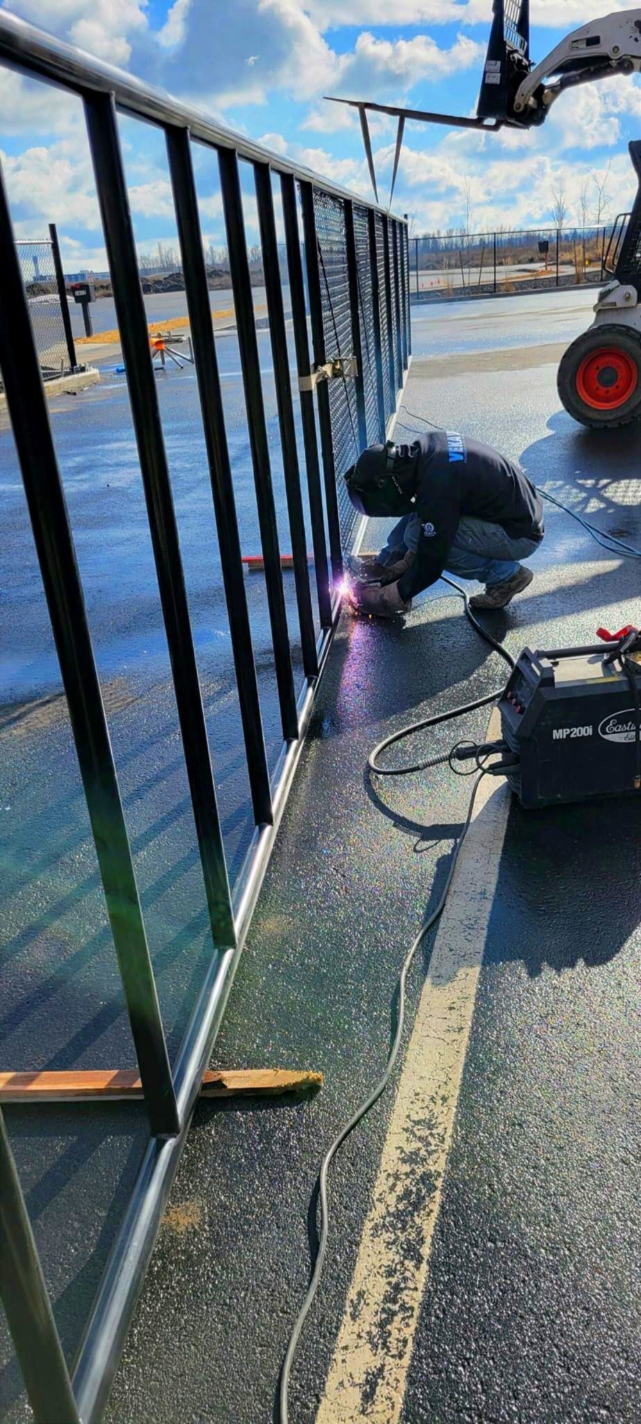 A worker welds a black metal fence on a wet asphalt surface next to a bobcat under a cloudy sky.
