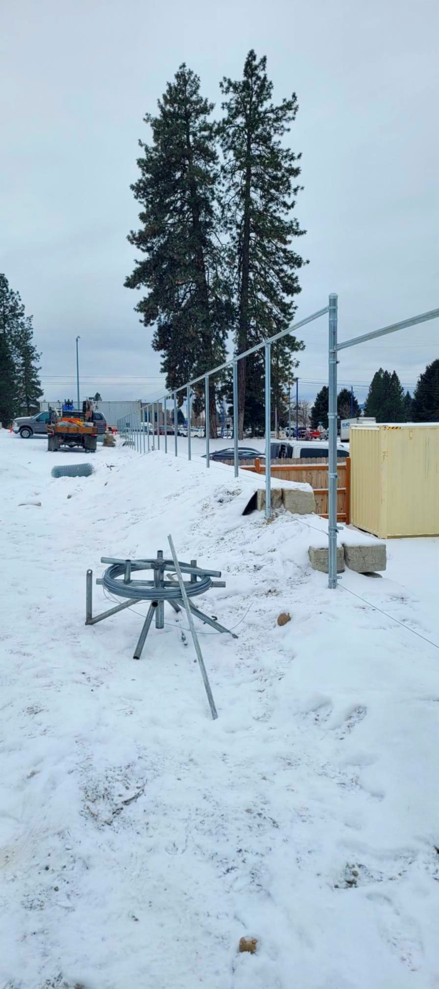 Construction site covered in snow, with a metal fence being built. Two tall trees are in the background.