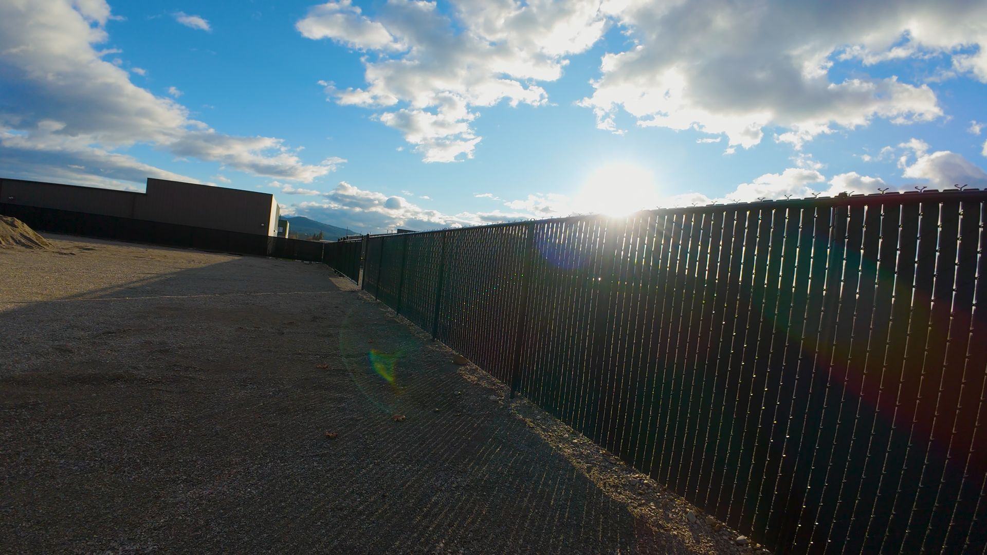 Sun shining through a dark privacy fence, with a blue sky and clouds overhead.