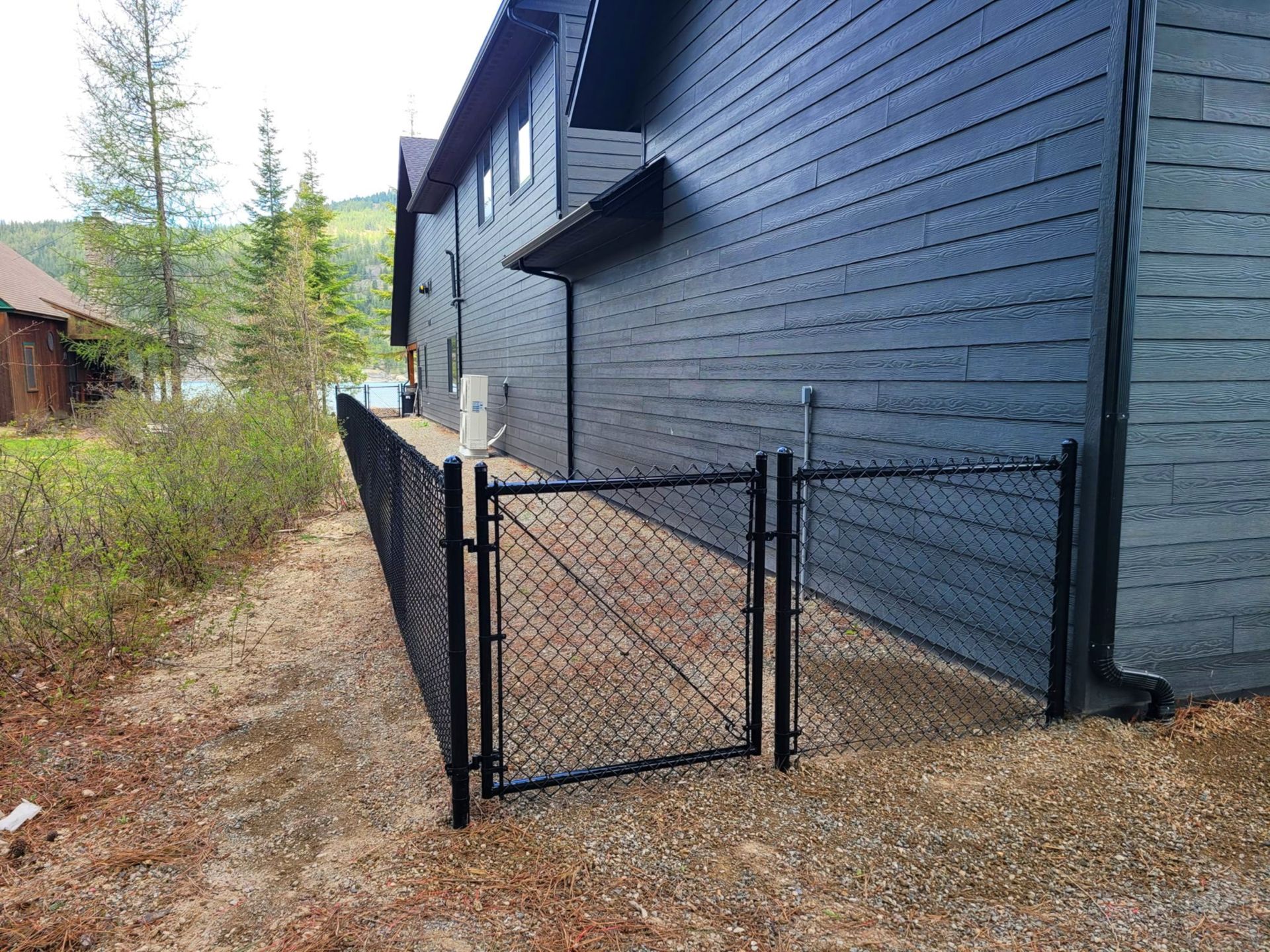 Black chain-link fence with gate beside dark blue house. Gravel ground, greenery, and trees in the background.