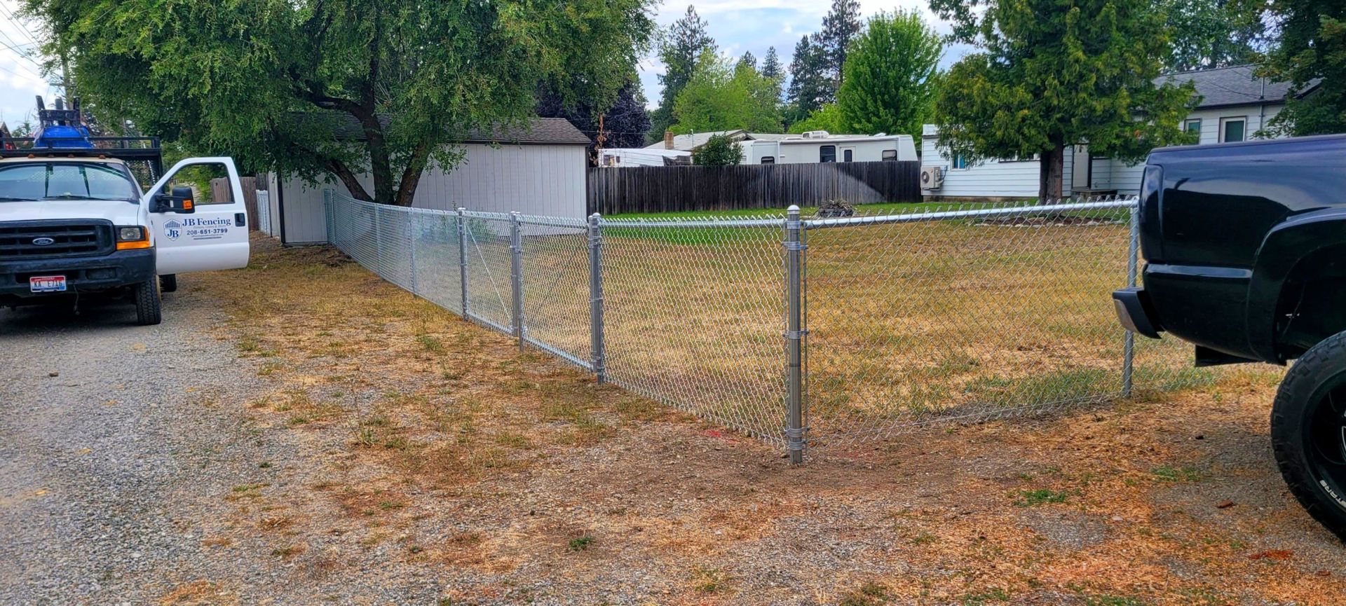 A chain-link fence lines a yard of dry grass next to a gravel driveway. A white truck and black truck are present.