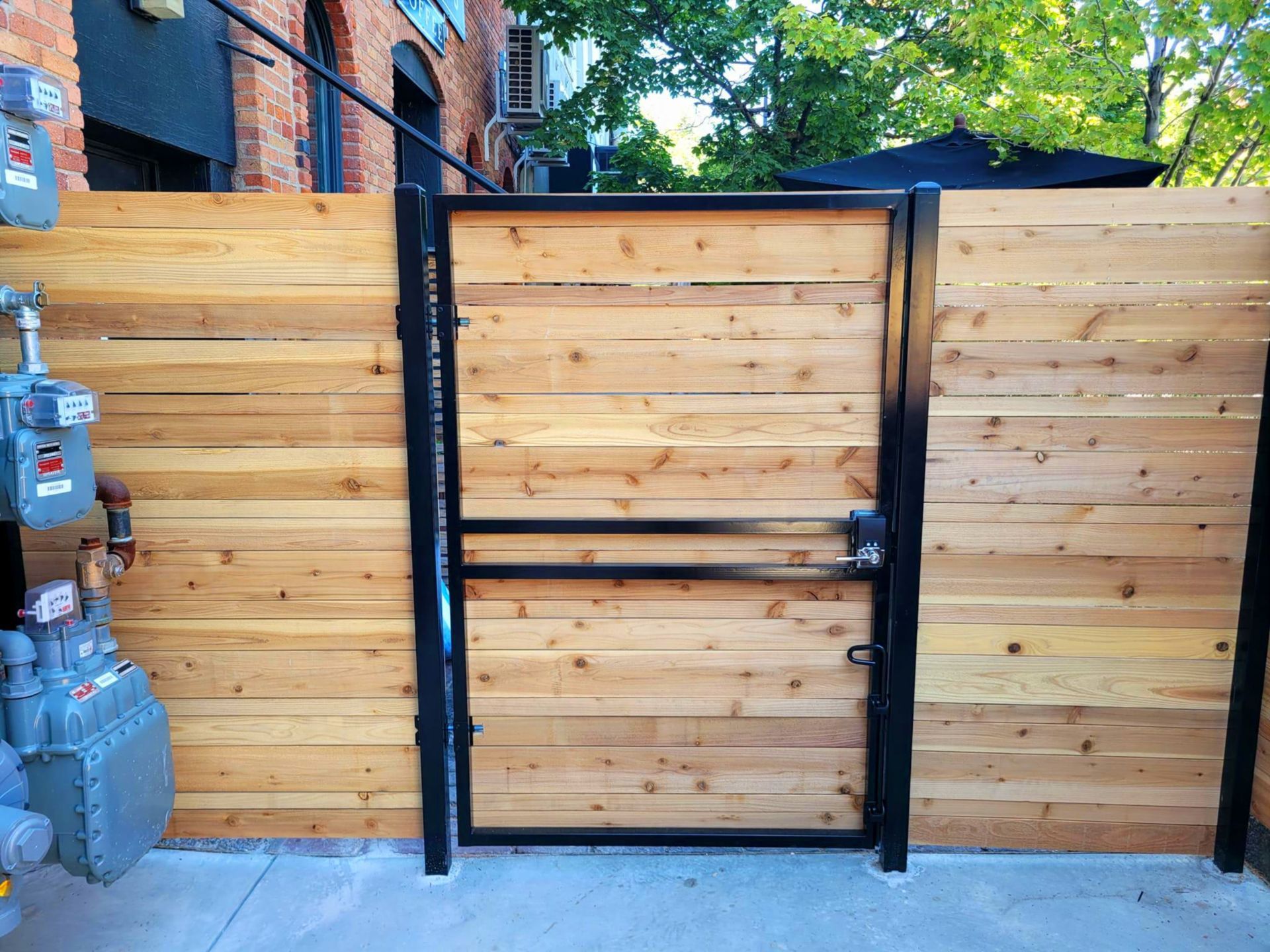 Wooden fence with black metal gate in front of a brick building.