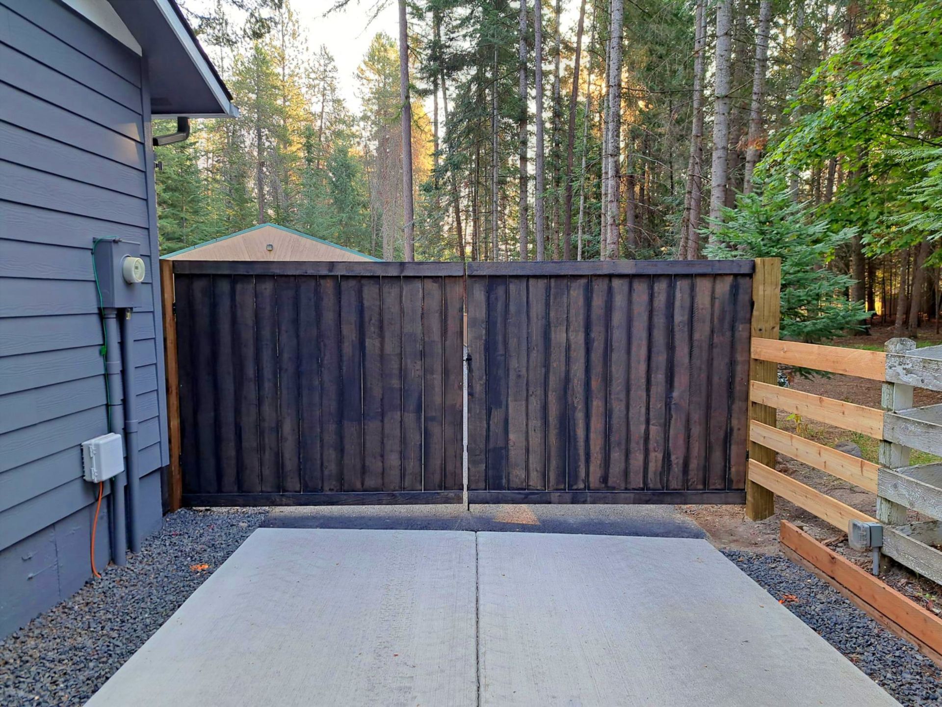 Dark wooden gate in backyard, concrete patio in front, gravel edging, forest background.