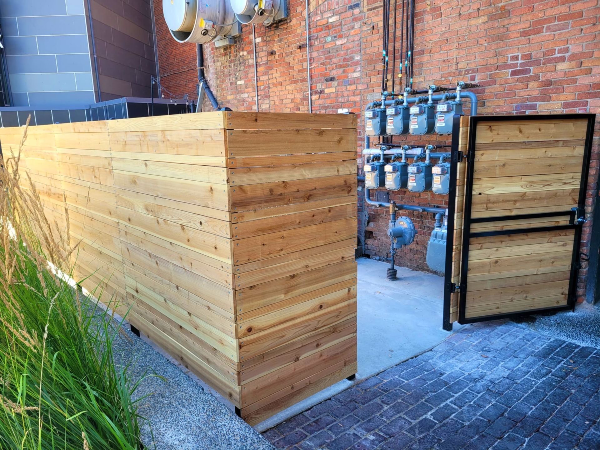 Wooden enclosure covering utility equipment next to a brick wall. A gate is open.