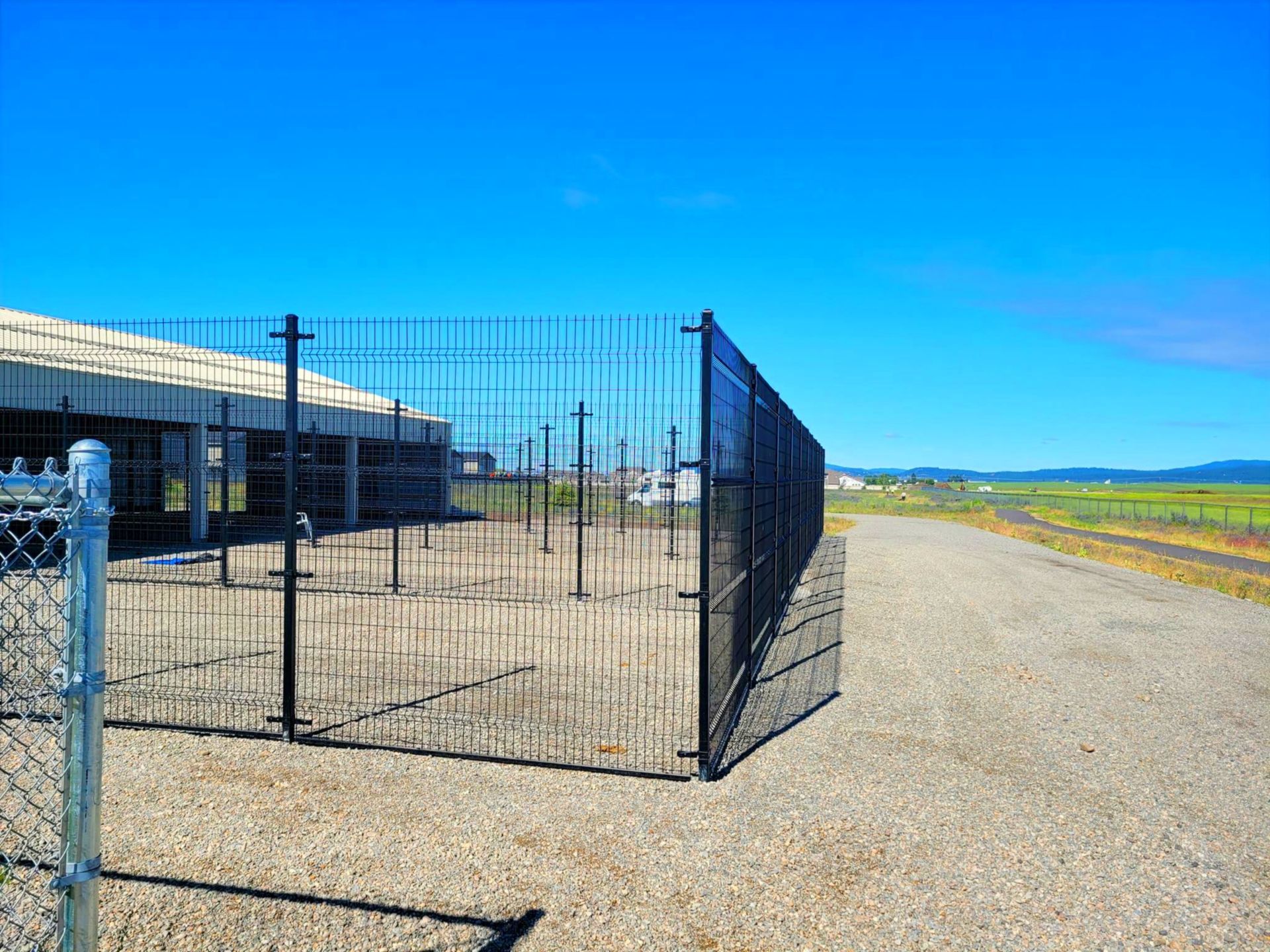 Black metal fence enclosing an area on a gravel path near a building and a marsh under a blue sky.