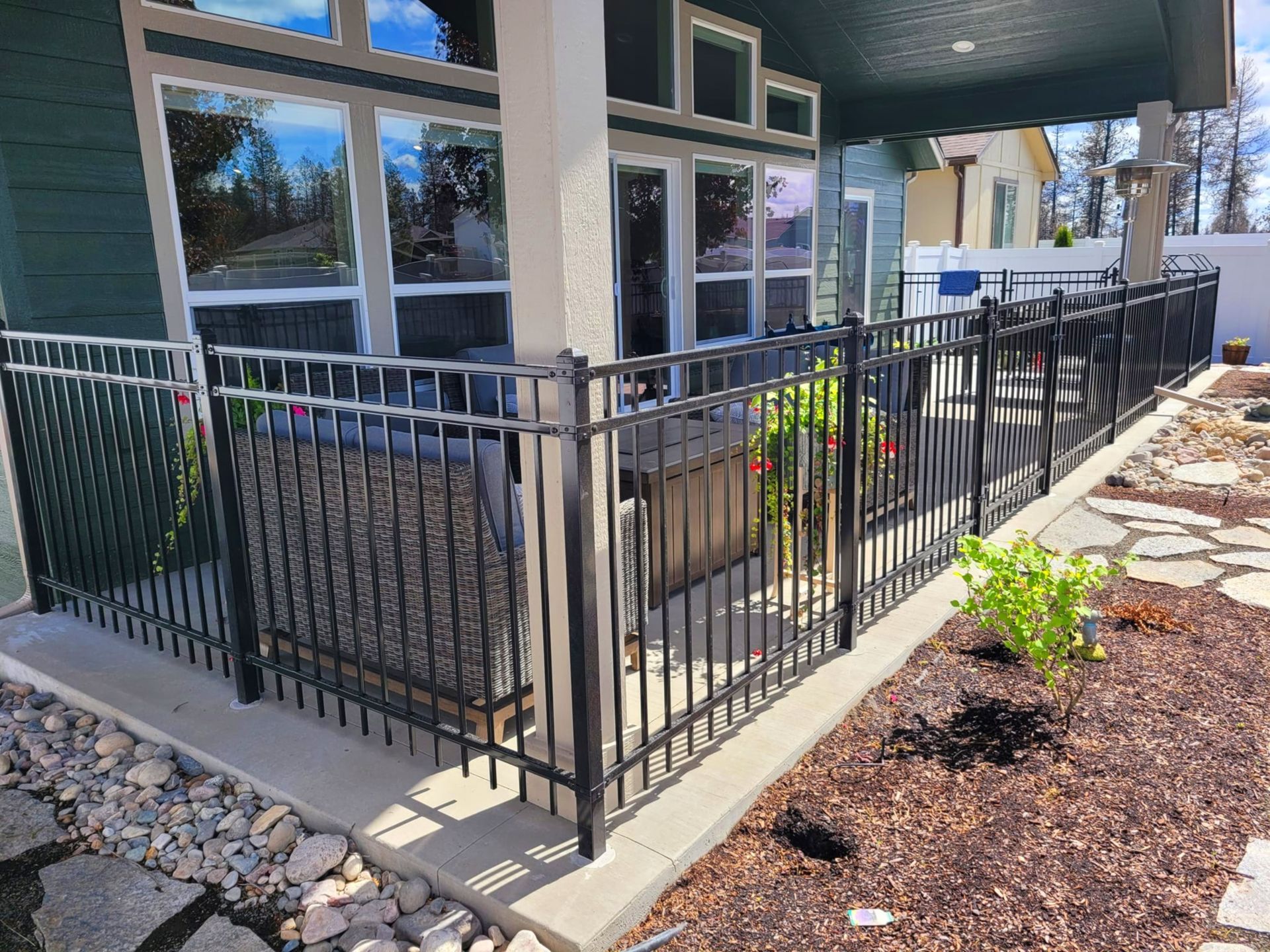 Black metal fence surrounds a patio with hot tub. Dark green house with large windows in background.