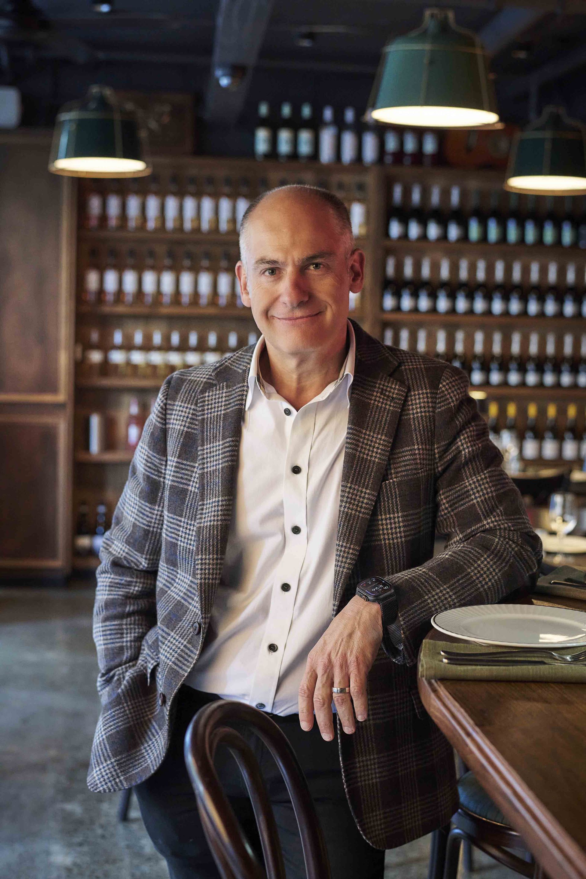 Man in plaid jacket smiles, leaning on a bar in a restaurant. Shelves of bottles in the background.