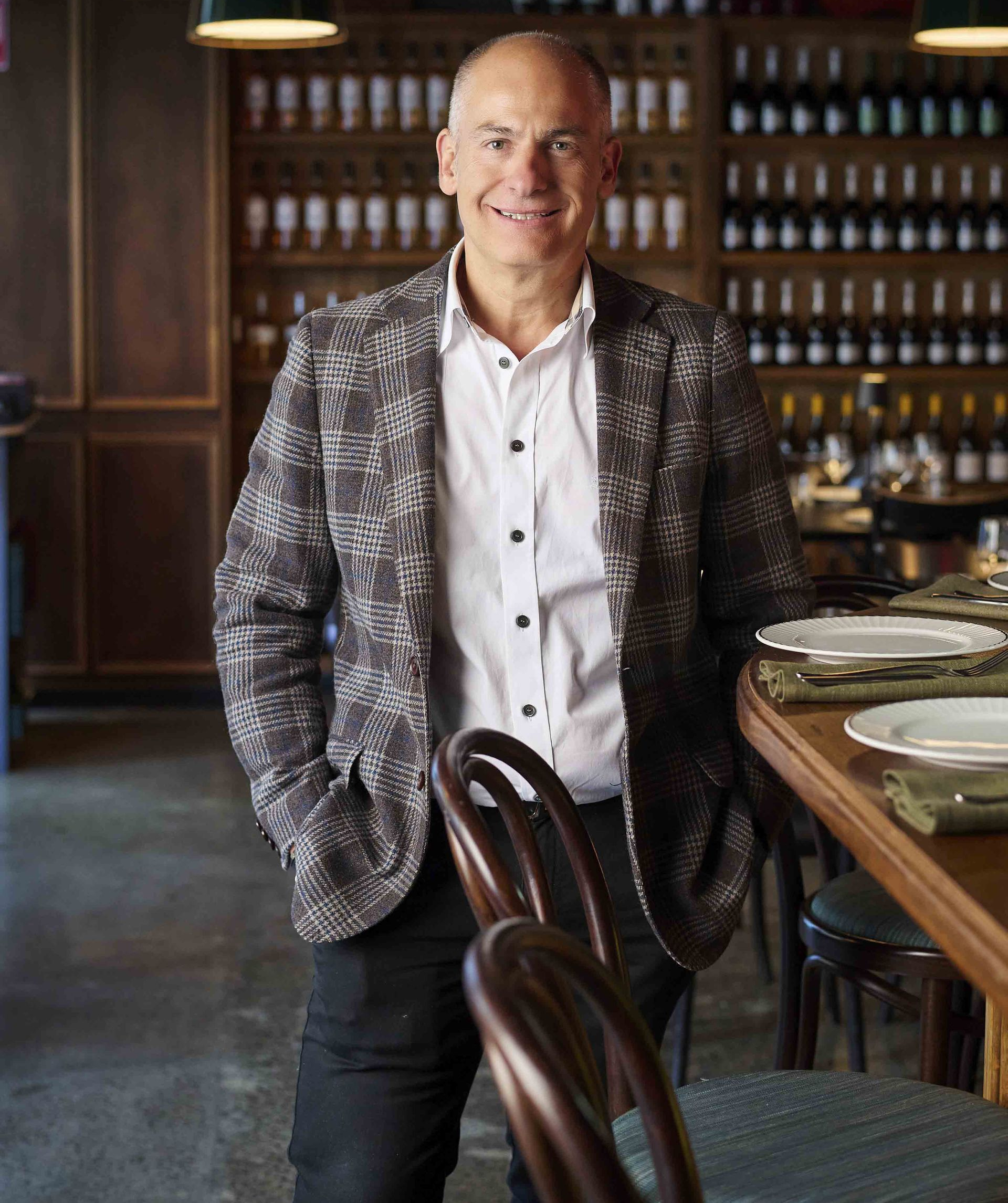 Man in patterned jacket, leaning on a bar, smiling. Restaurant interior with wine bottles visible.