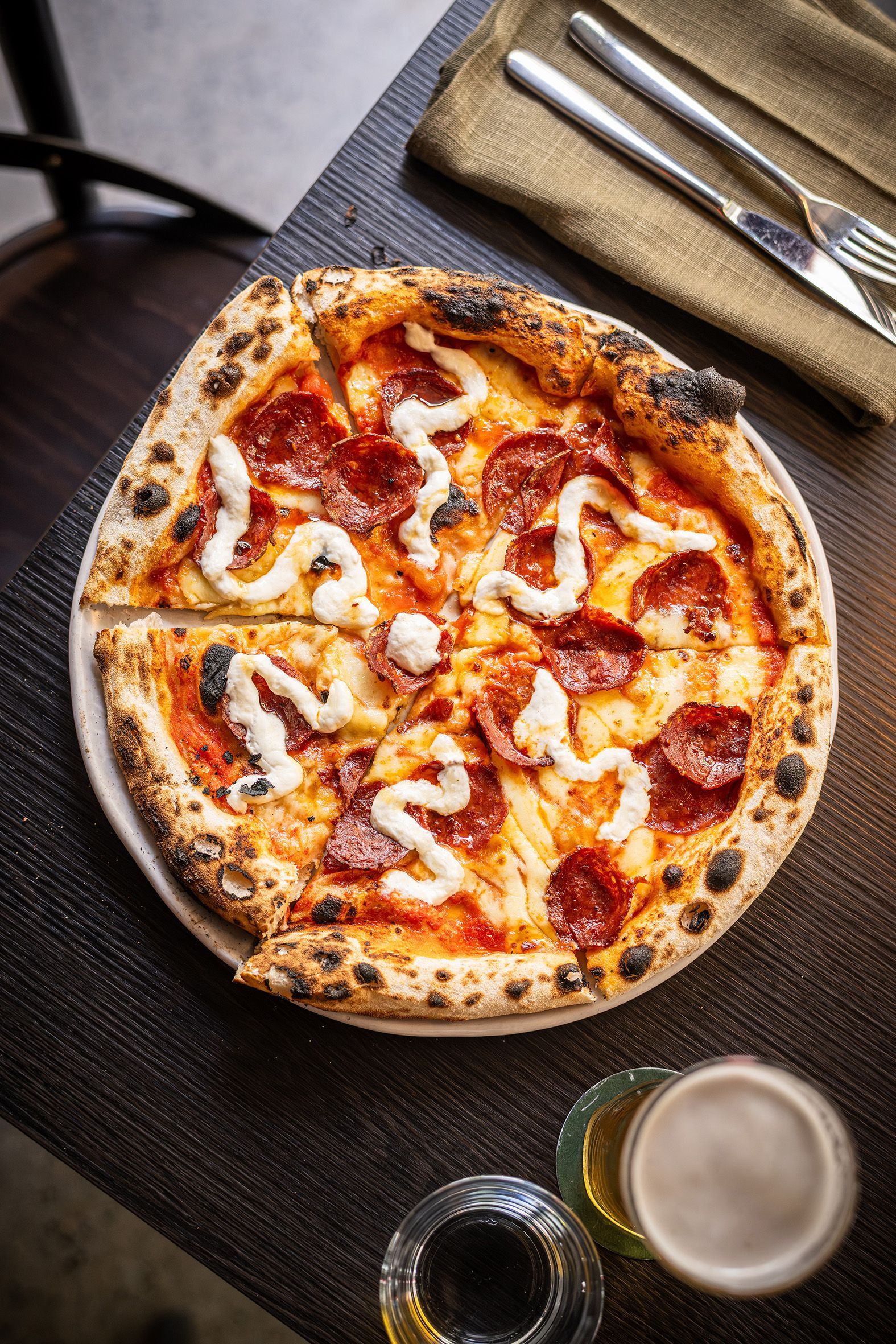 Pepperoni pizza with white sauce, on a plate, next to silverware on a burlap napkin, with a drink on a table.