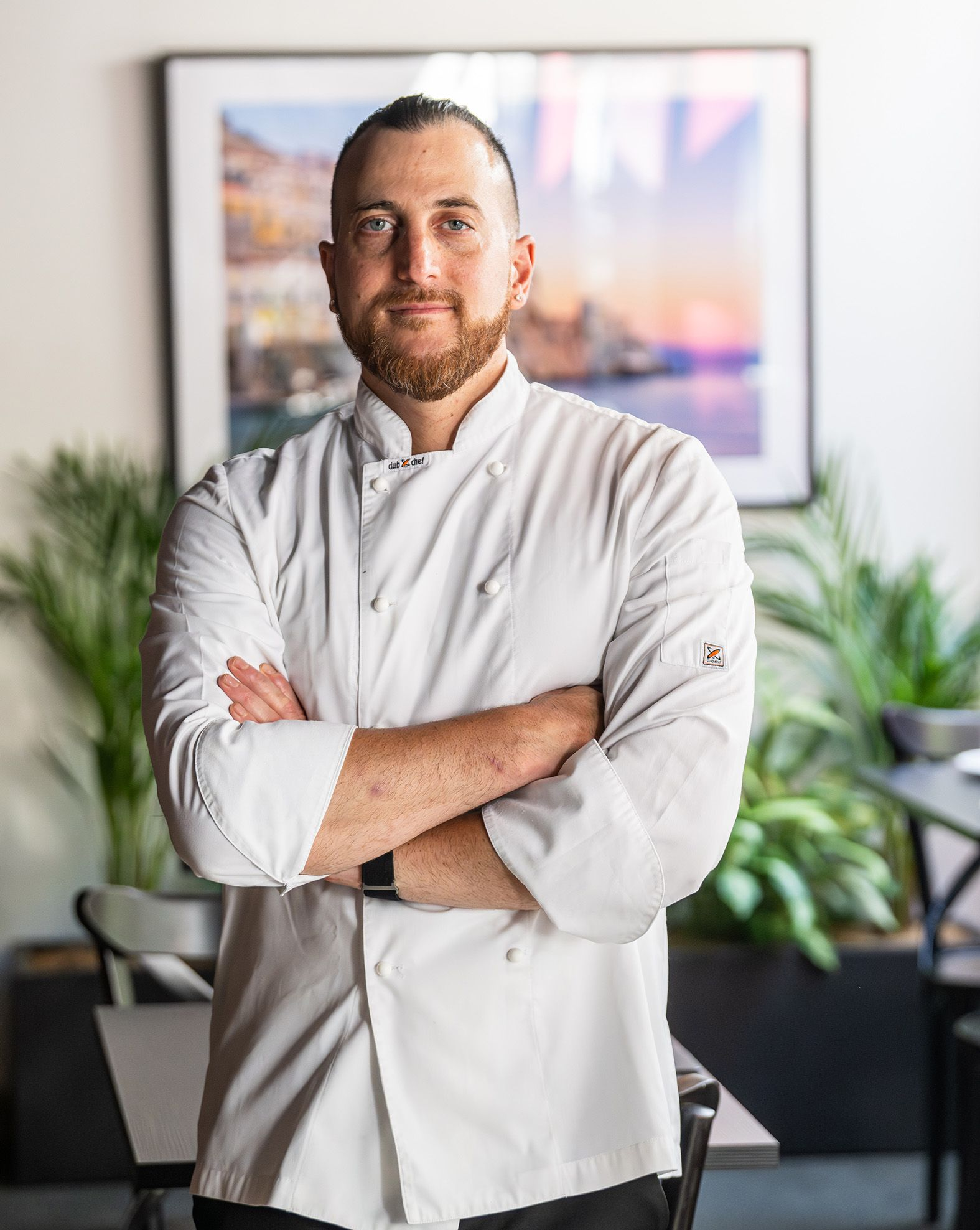 Chef in white coat, arms crossed, standing in a restaurant.