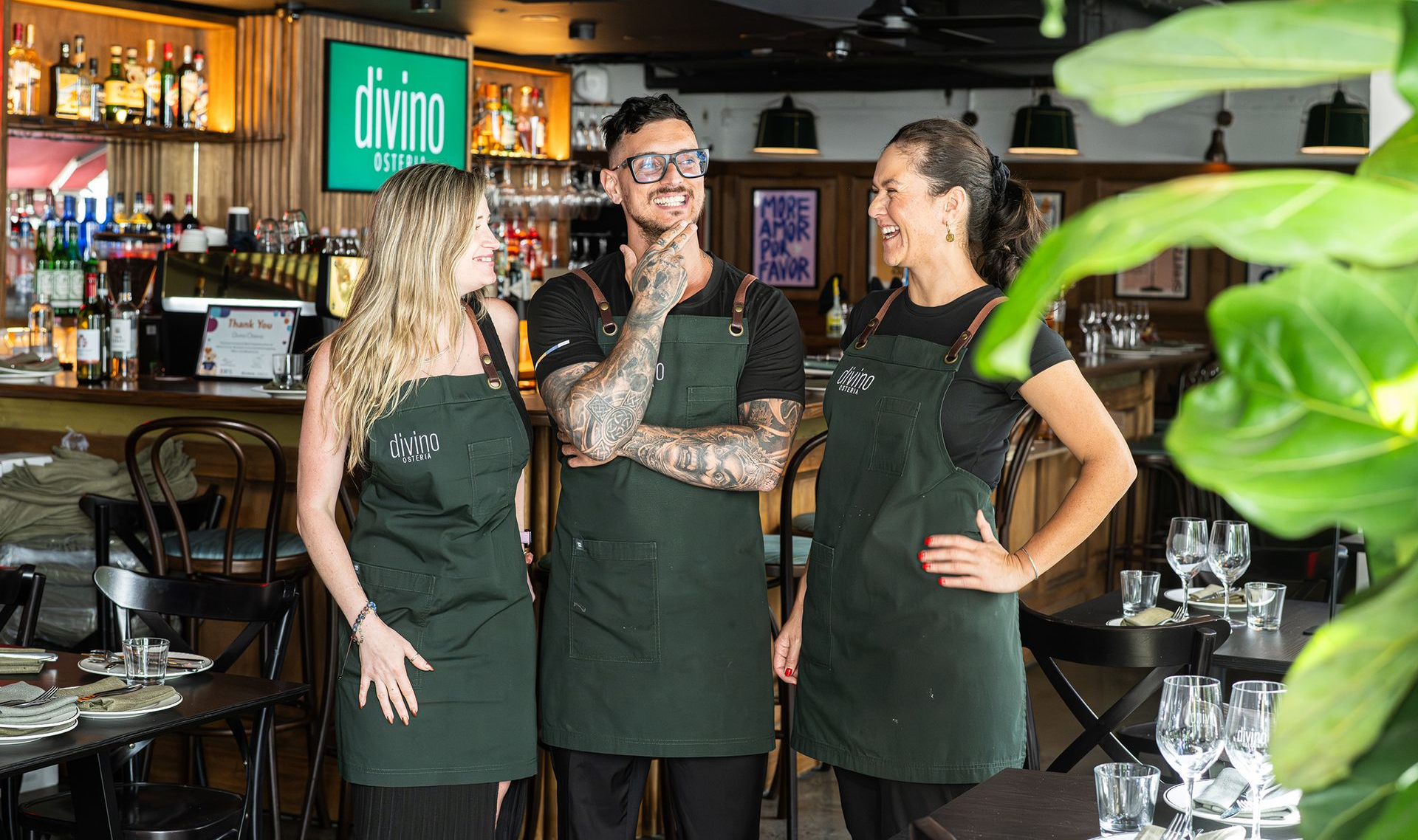 Three restaurant staff, wearing green aprons, pose smiling inside; bar with drinks visible behind them.