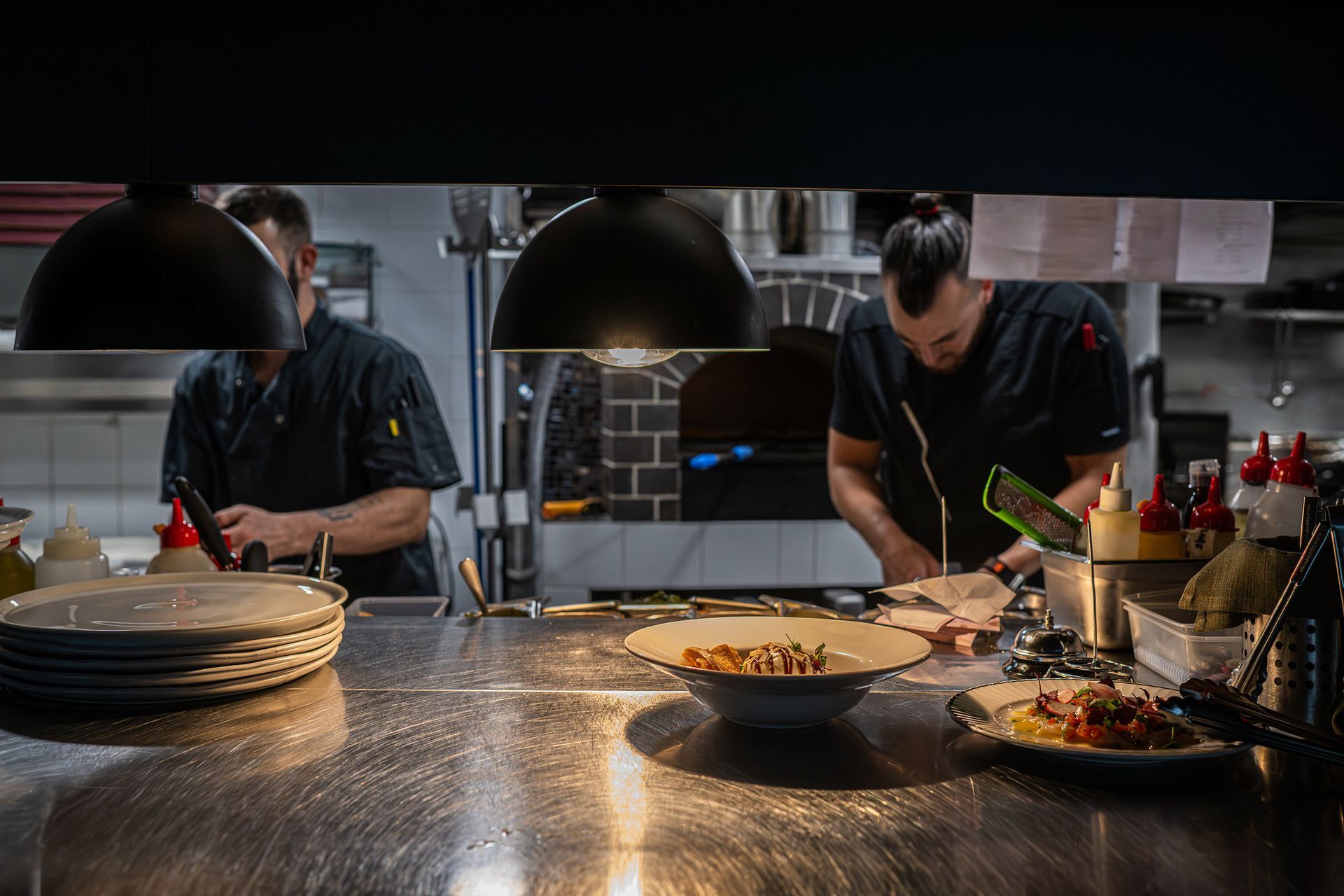 Two chefs working in a commercial kitchen with plates and food on a stainless steel counter.