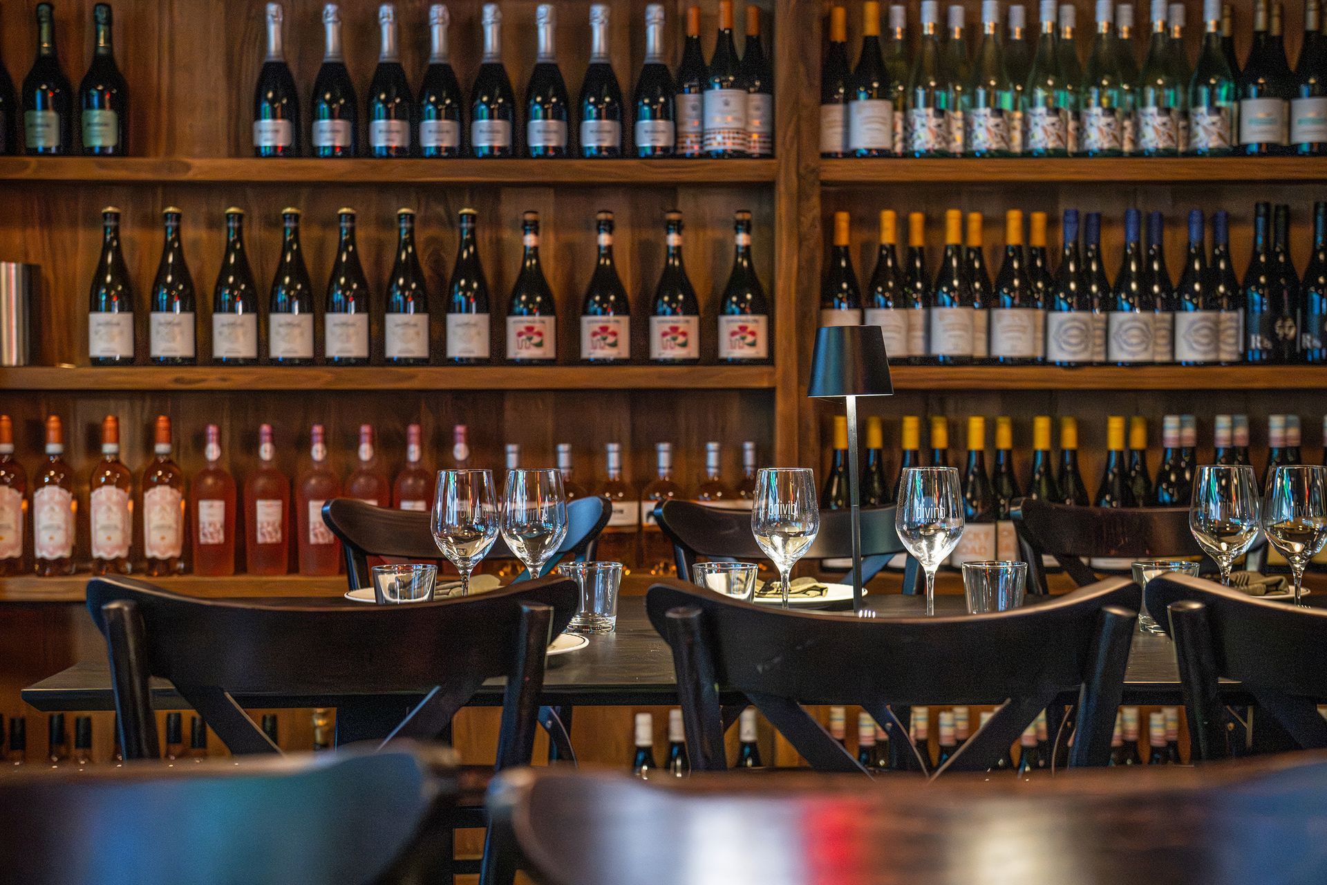 Restaurant interior with wooden shelves filled with wine bottles; tables with glasses are set in front.