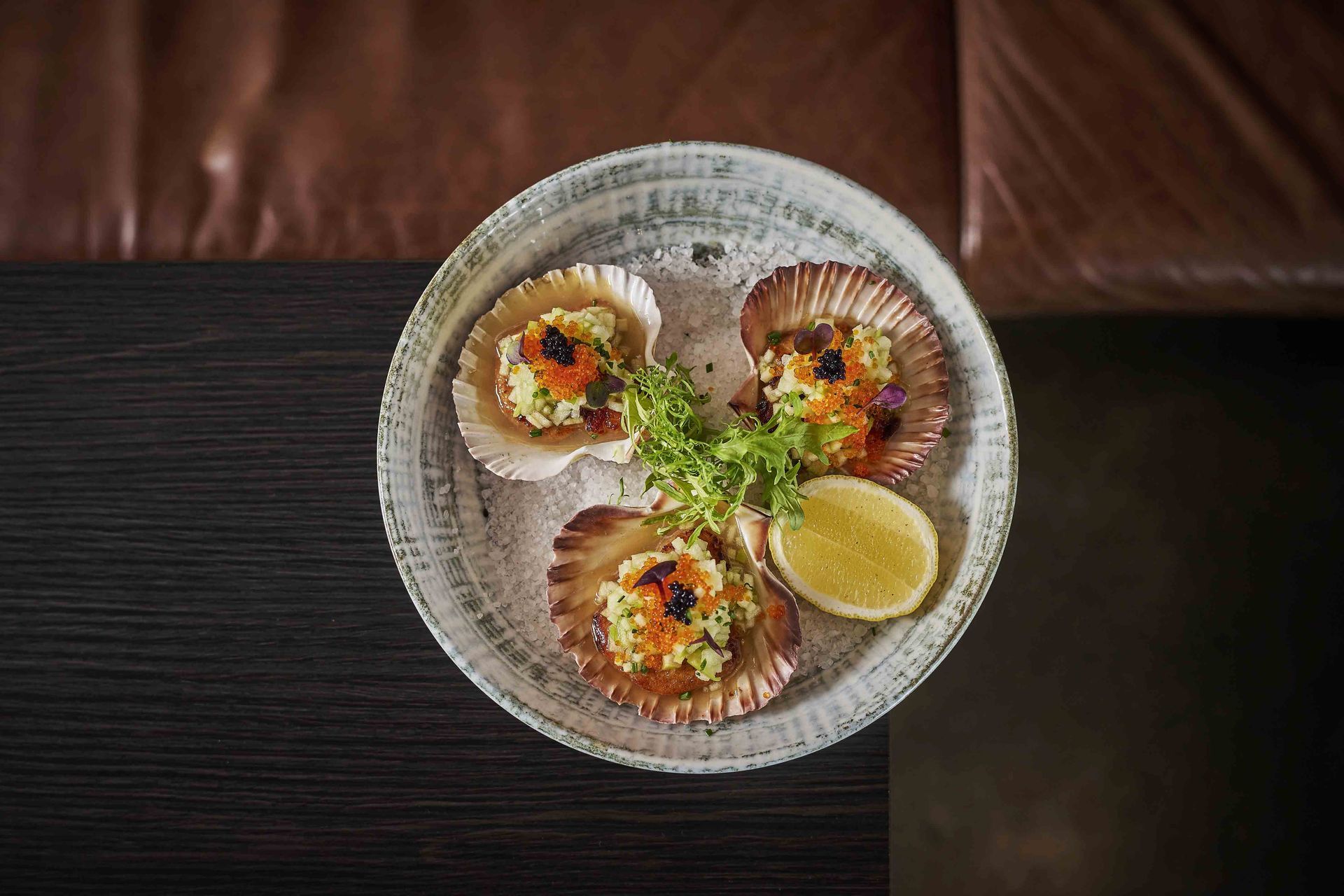 Seared scallops on shells, topped with green mixture and caviar, served on a white plate with lemon.