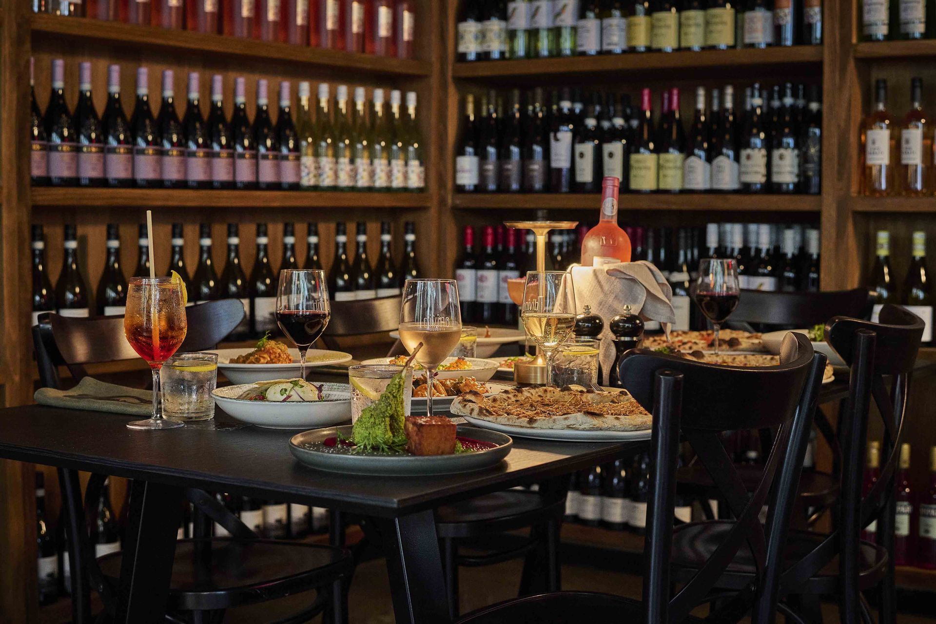Table set with food and drinks in front of wine-filled shelves.