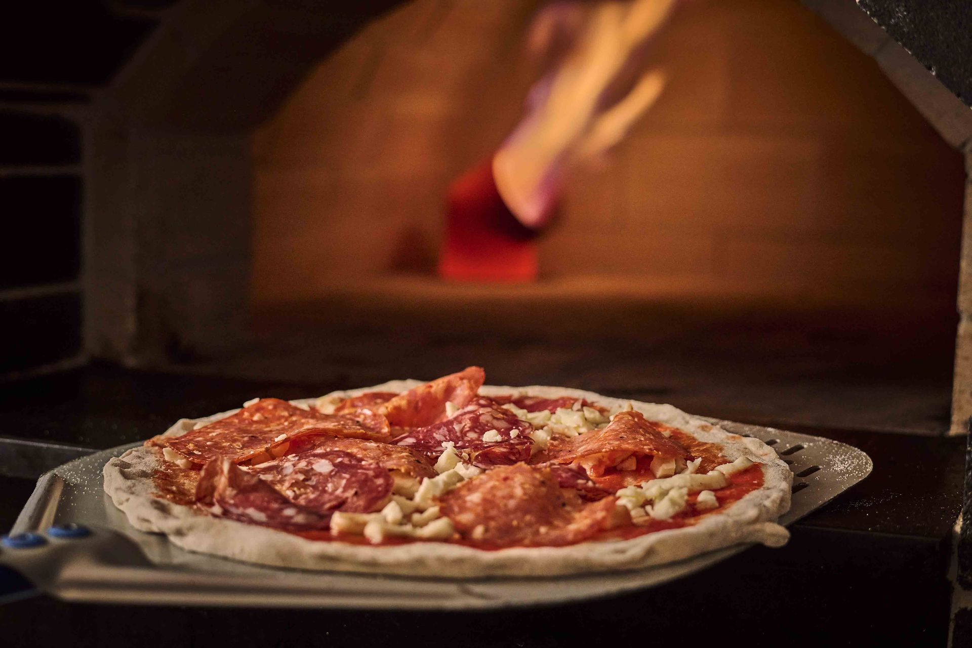 Pizza on a peel being placed into a brick oven with flames visible in the background.