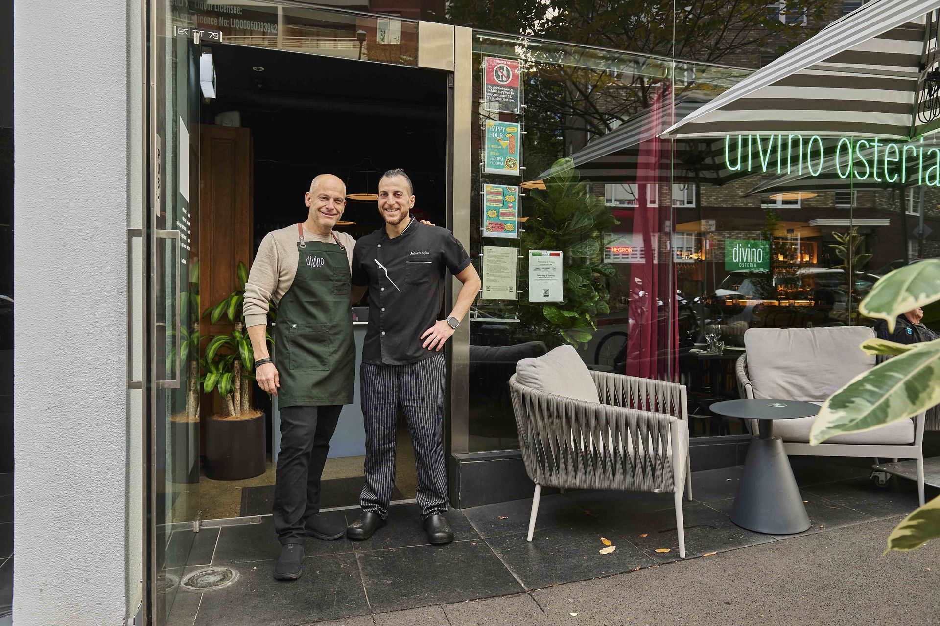 Two men stand in front of Divino Ostaria, an outdoor restaurant. One wears an apron, the other a chef's uniform.