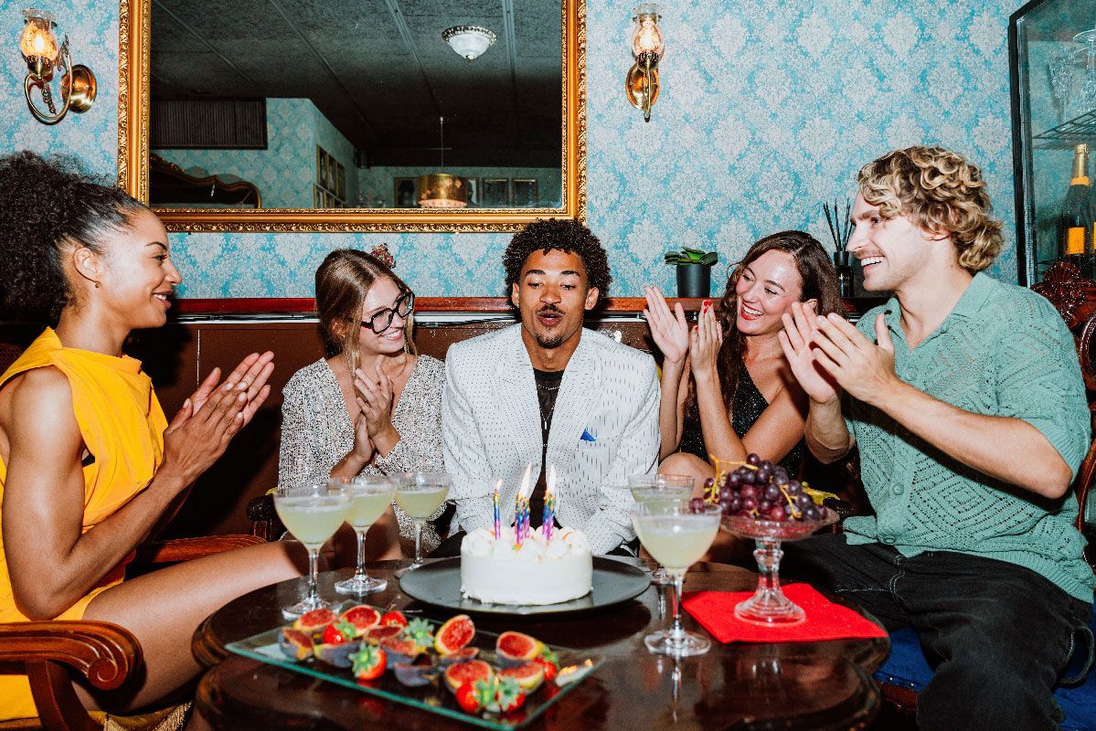 Friends celebrate a birthday with cake and drinks in a room with blue patterned wallpaper. People clap.