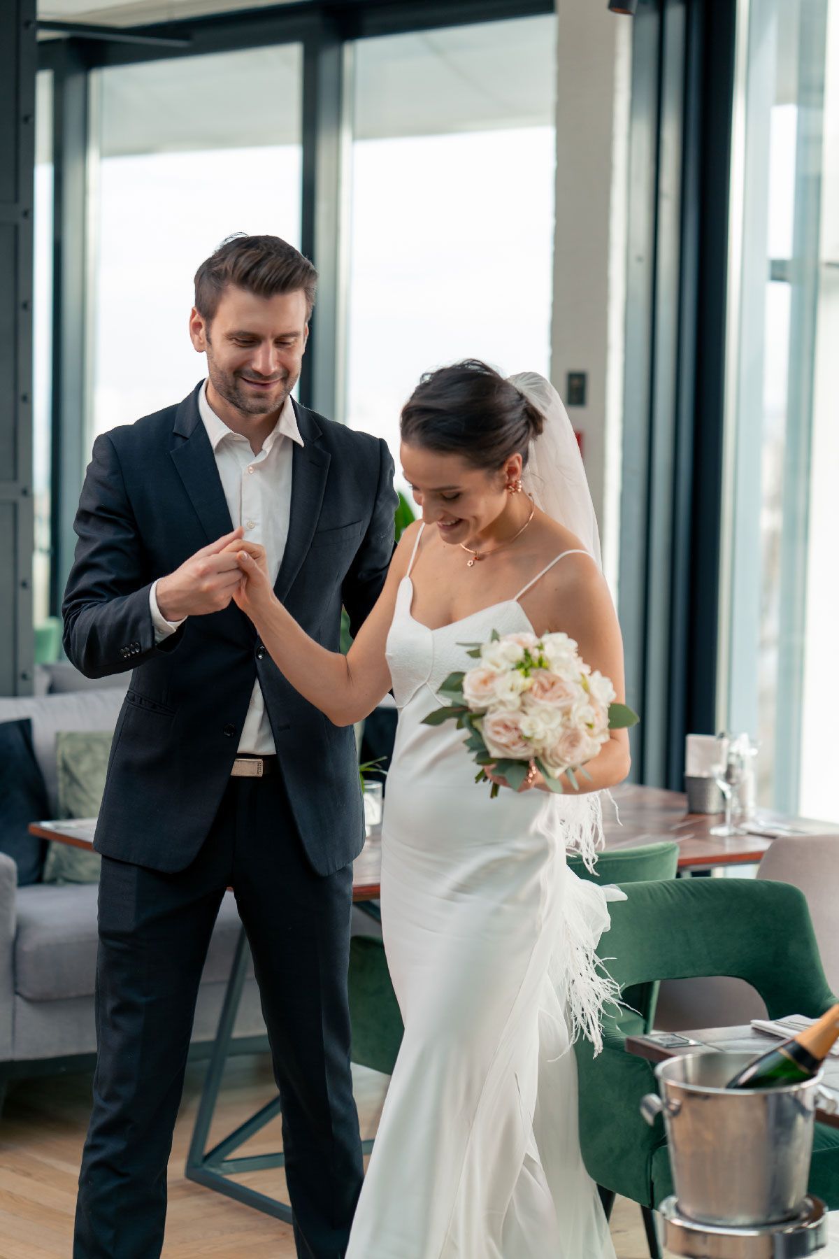 Bride and groom holding hands, smiling. She wears a white gown, he a dark suit, near a table.