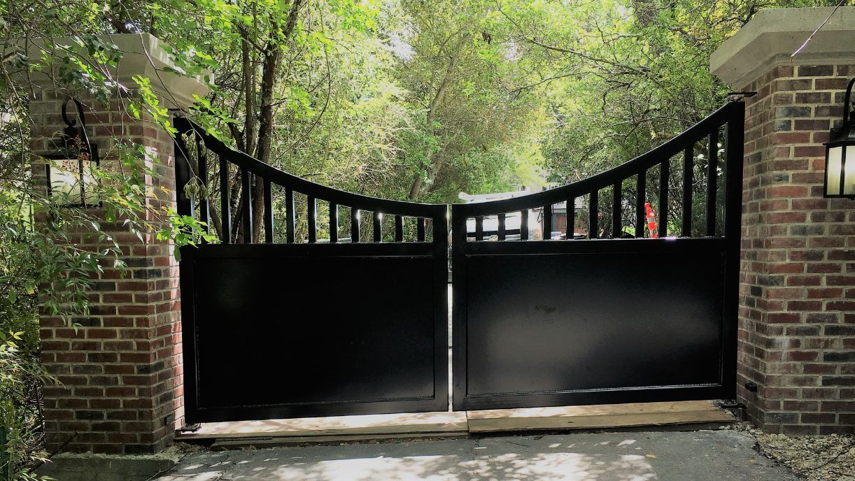 A black gate is open to a driveway surrounded by trees.
