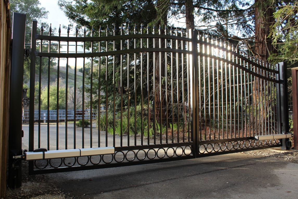 A wrought iron gate is open to a driveway with trees in the background.