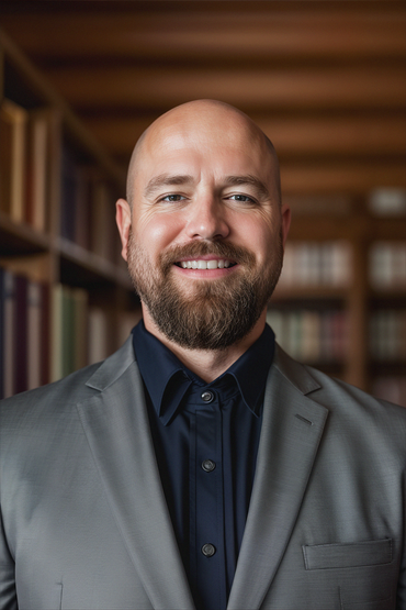 Man in gray suit and dark shirt smiles in library, books in background.