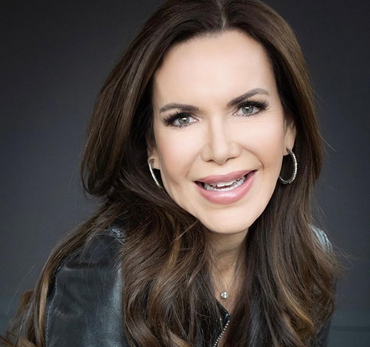 Woman with long brown hair, smiling, wearing a black leather jacket and hoop earrings.