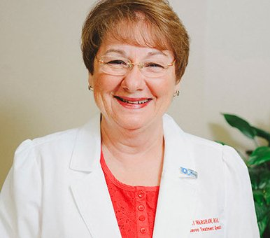 Woman wearing glasses and a white coat, smiling, inside.
