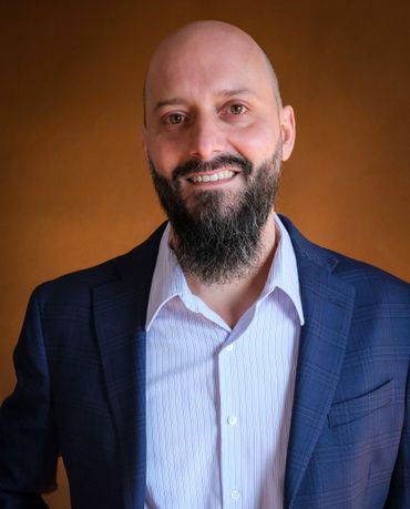 Man with a bald head and beard wearing a blazer, smiling in front of a brown backdrop.
