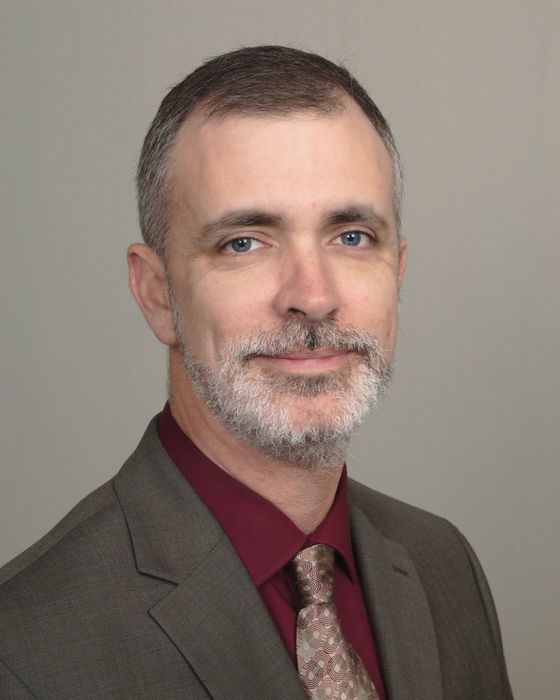 Man in a suit, grey hair and beard, wearing a red shirt and patterned tie, looking at the camera.