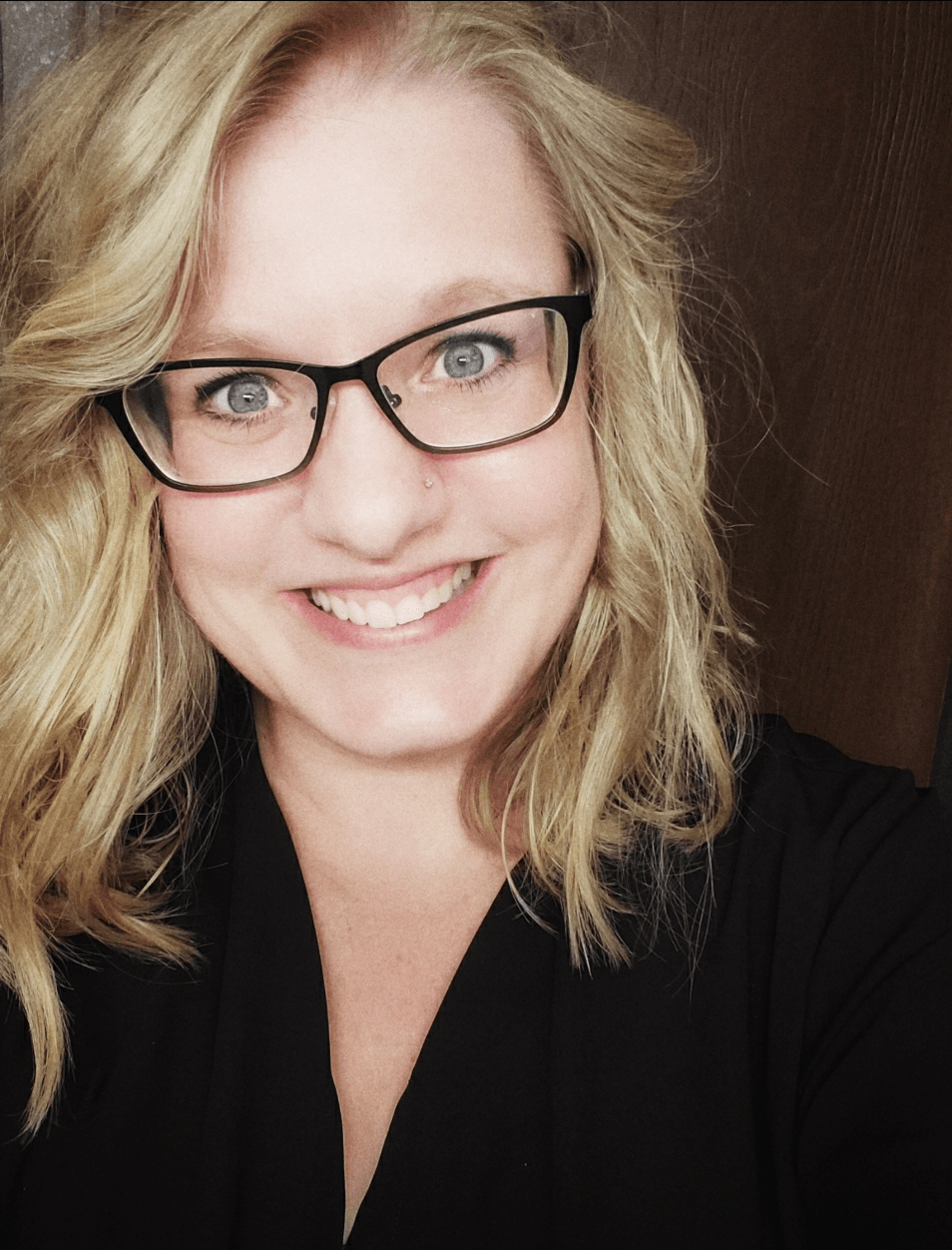 Woman with blonde hair and glasses smiles broadly, wearing a black shirt, indoors.