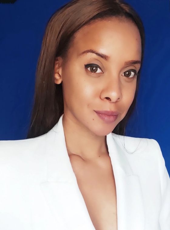 Woman in white blazer, brown hair, looking at the camera against a blue backdrop.