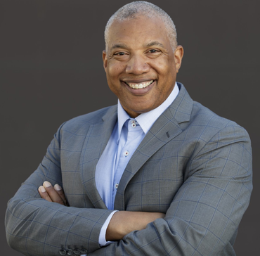 Man in a gray suit with arms crossed, smiling broadly against a gray background.