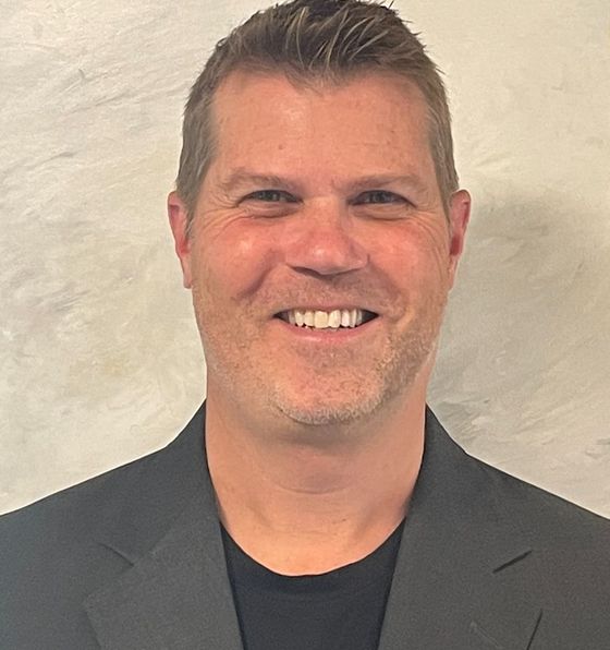 Man with short hair and a broad smile wearing a black shirt and gray jacket.