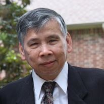 Man in a suit smiles, gray hair, outdoors near a brick building.