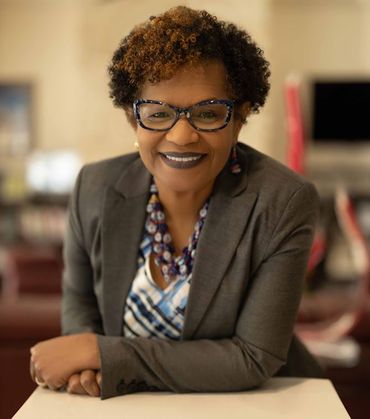Woman in glasses and gray blazer smiles, leaning on a white surface.