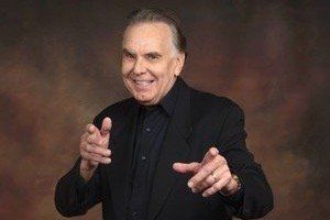 Smiling man in black blazer points towards the viewer with both hands, against a brown backdrop.
