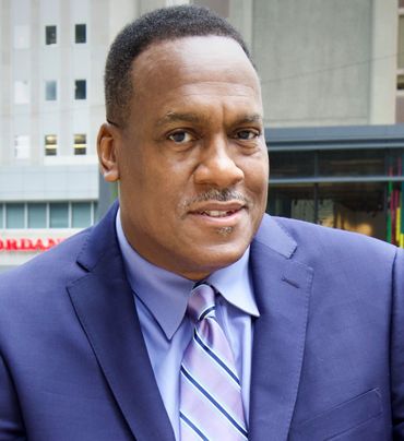 Man in a blue suit and purple tie smiles, outdoors with a city backdrop.