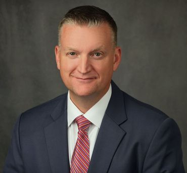 Man in a navy suit and red striped tie, smiling, against a dark gray backdrop.