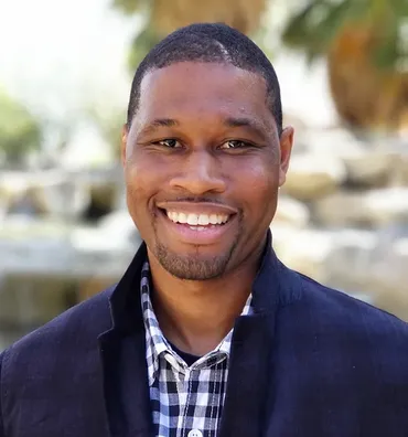 Man with dark skin smiling, wearing a blue blazer over a checkered shirt, in a sunny outdoor setting.