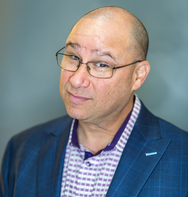 Man with glasses and shaved head, wearing a patterned shirt and blue blazer, looking at the viewer.