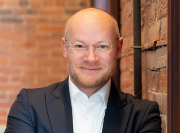 Man in a dark suit and glasses smiles, leaning against a brick wall.